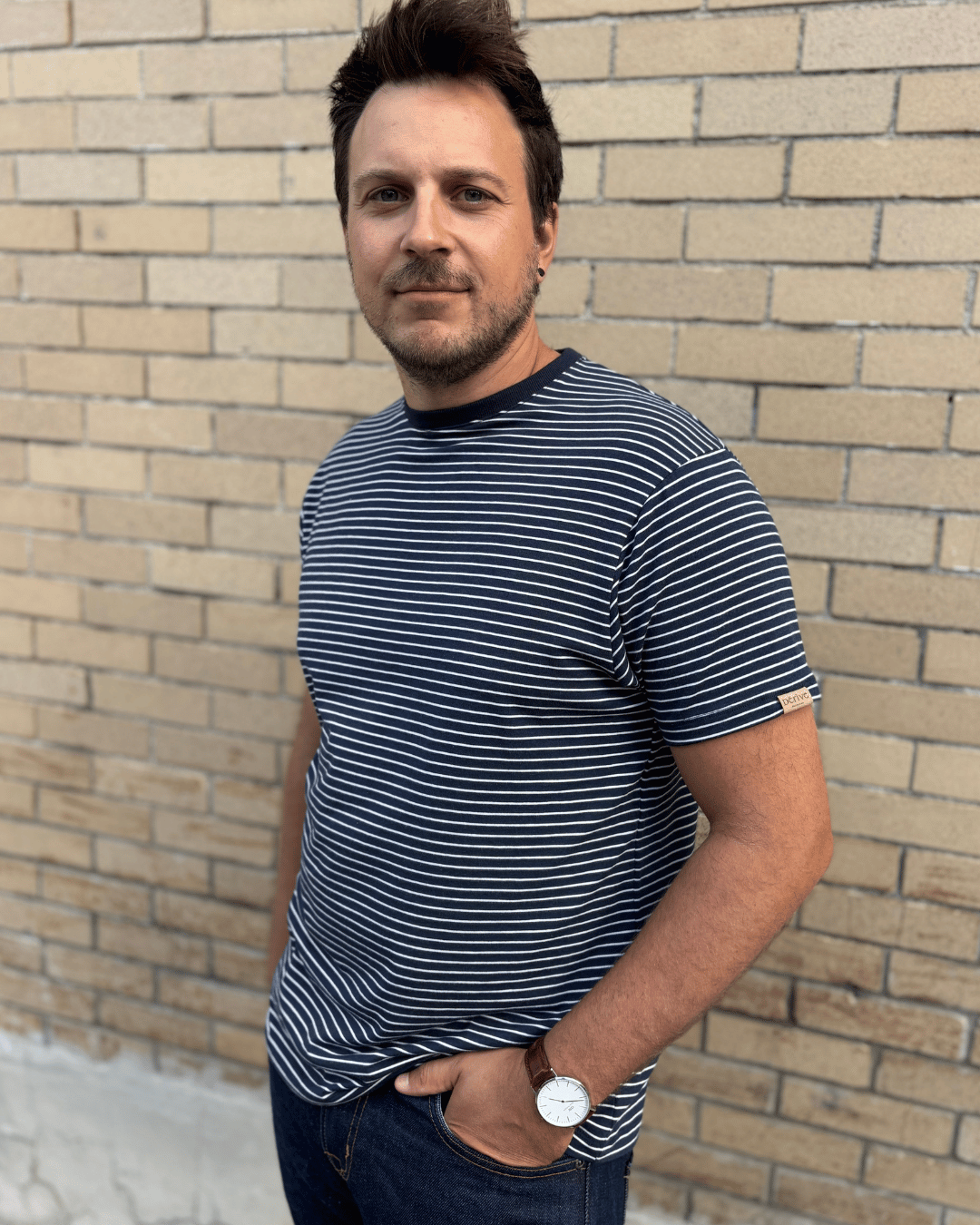 Homme portant un t-shirt à manches courtes rayé bleu marine et blanc avec jeans foncés et montre au poignet, debout devant un mur de briques beiges.