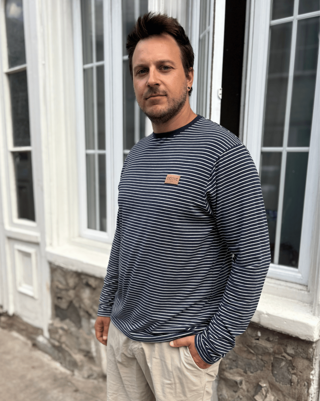 Homme portant un t-shirt à manches longues rayé noir et blanc avec un pantalon beige devant une façade en pierre et fenêtres blanches