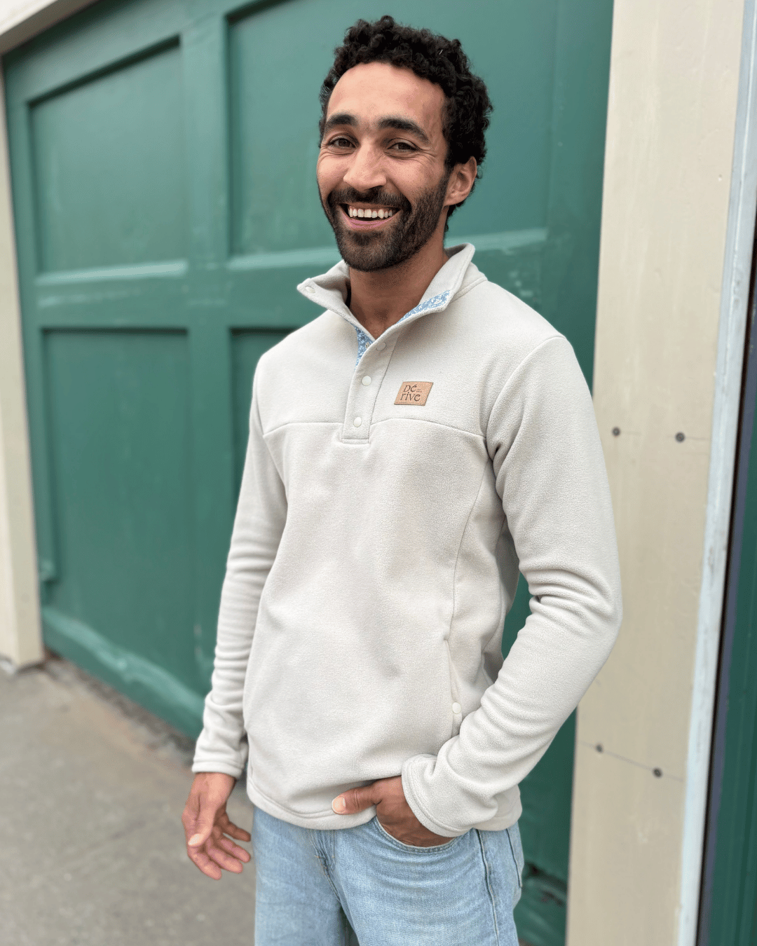 Homme souriant debout à l’extérieur, portant un pull beige clair et un jean devant une porte verte.